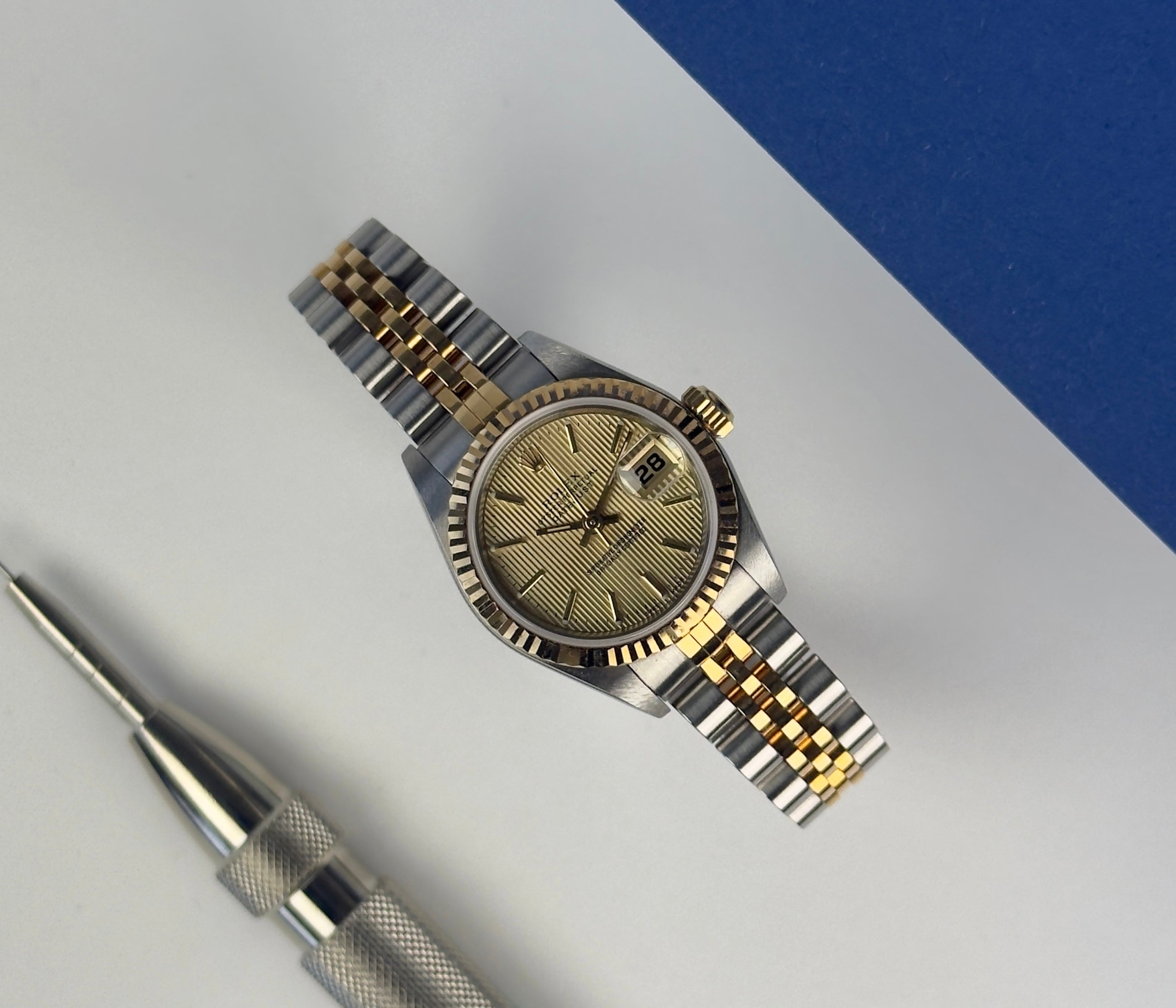 Rolex Datejust 79173 (2002) i guld og stål. Dette klassiske dame Datejust i 26 mm har en champagne Tapestry skive, jubileelænke og safirglas. Lænke uden stræk. Uret fremstår i rigtig flot stand, 9/10. Et utrolig flot og feminint ur, der er den perfekte kombination af ur og smykke.