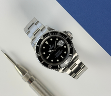 Rolex Submariner 16610 T V-serie (2009) solgt i 2010. Uret fremstår i flot stand, 9/10. Det måler 40 mm og er monteret på en Oysterlænke med sort aluminiumslynette. Kommer med bokse og certifikat.